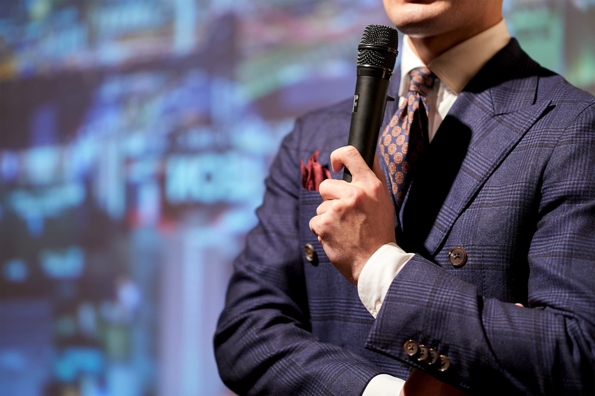 スーツを着た人物がマイクを持ち、スクリーンを背にプレゼンテーションを行っている場面。